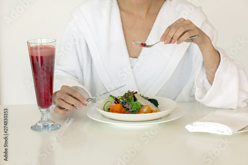 Fresh tasty vegetable salad berry drink and woman in white fluffy robe eating with knife and fork at table