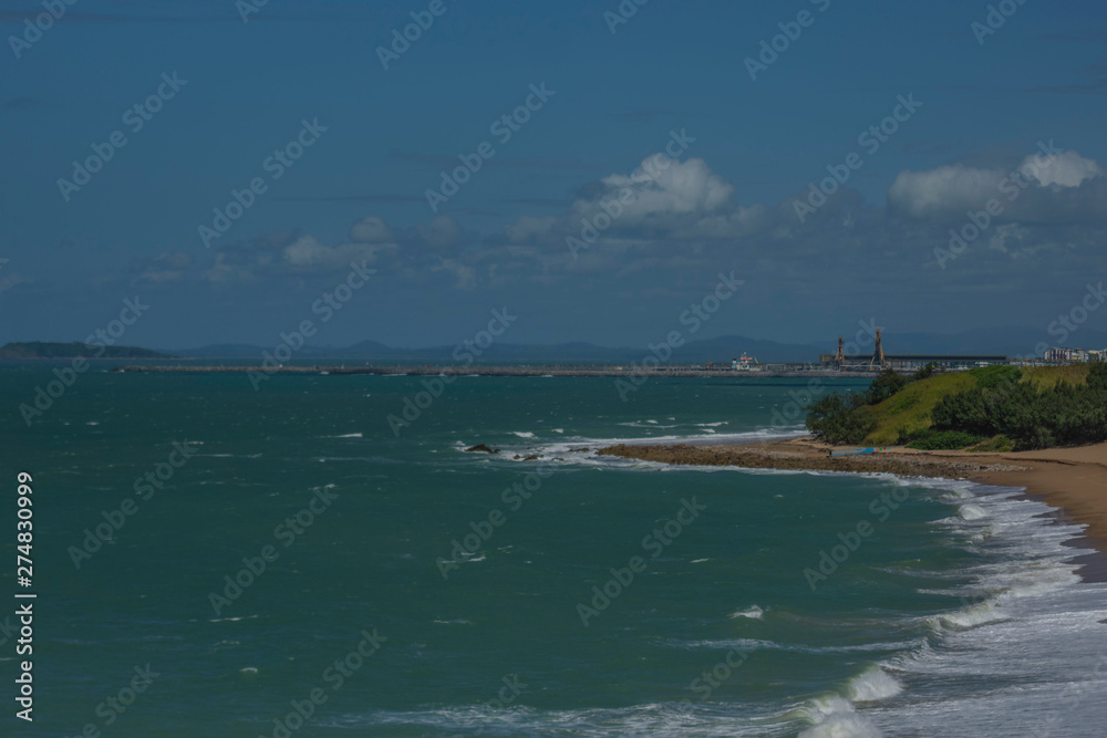 Sandy beach with gentle sea waves  and industrial towers in background against blue sky