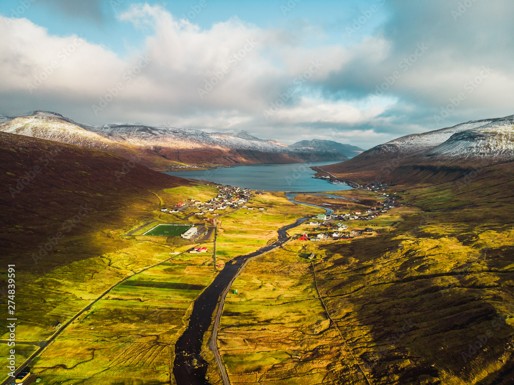 Plakat Aerial photography of Faroese village Streymnes during sunset in spring with cloudy blue sky, sunny green fields and snowy mountains (Faroe Islands, Denmark, Europe)