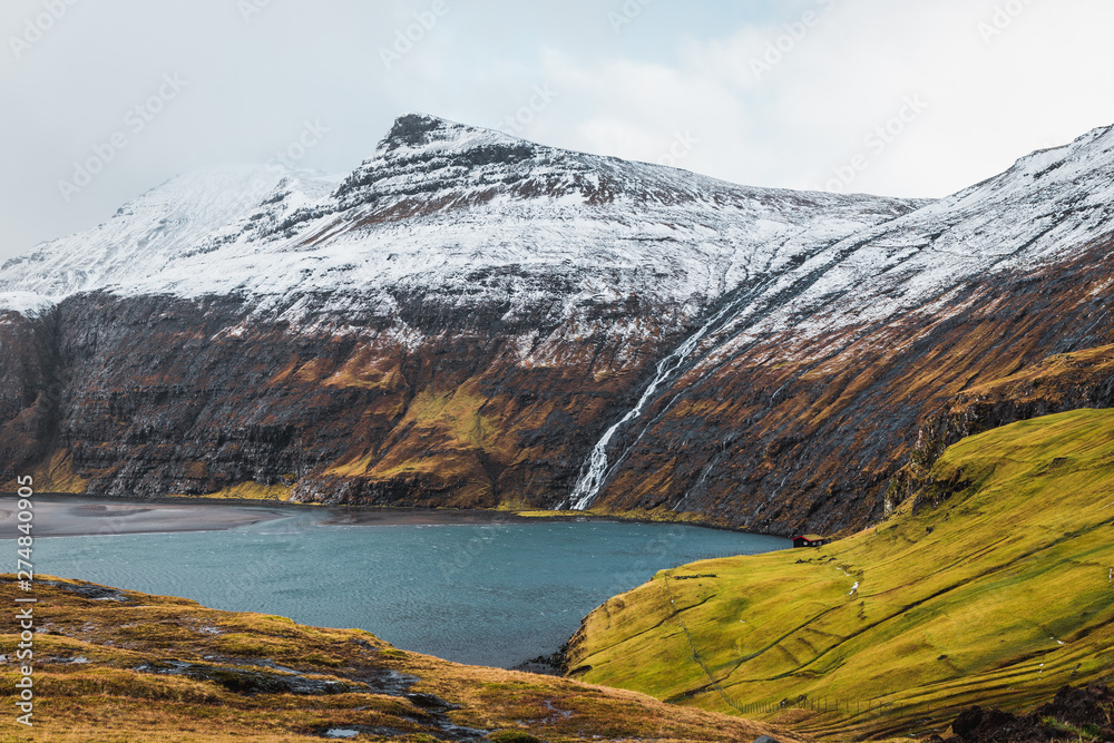 Lonely cabin in the fjord of Saksun during early spring with snow-covered mountains, a waterfall and lush green and orange grass (Faroe Islands, Denmark, Europe)