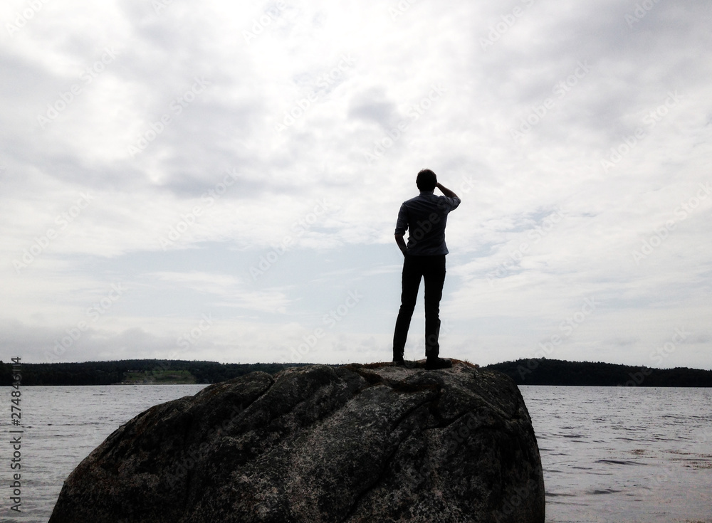 Silhouette of man standing on large rock looking out into a lake