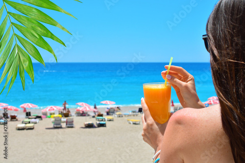 Fototapeta Naklejka Na Ścianę i Meble -  Girl holds orange fresh juice in her hand. Blue sea and beach on a backdrop.