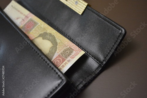 Black leather wallet with money in it on a brown matte background. Banknote 100 one hundred Ukrainian hryvnia inside a black wallet.