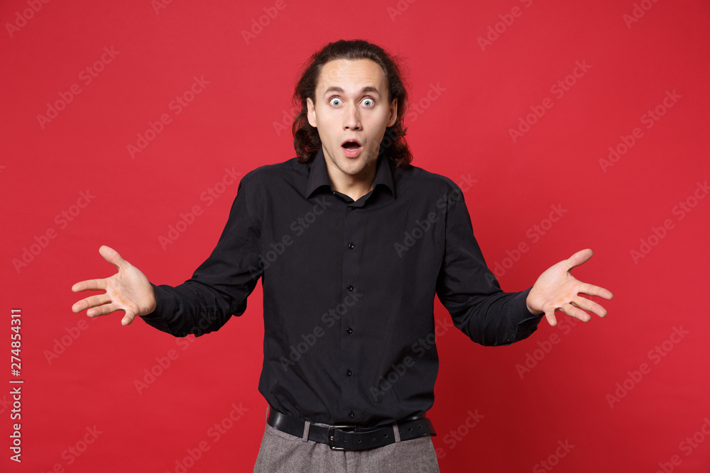 Shocked upset stylish young curly long haired man in black shirt posing isolated on red wall background studio portrait. People sincere emotions lifestyle concept. Mock up copy space. Looking camera. Shocked upset stylish young curly long haired man in black shirt posing isolated on red wall background studio portrait. People sincere emotions lifestyle concept. Mock up copy space. Looking camera.