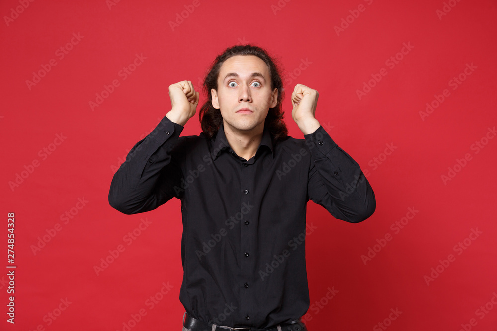 Shocked upset stylish young curly long haired man in black shirt posing isolated on red wall background studio portrait. People sincere emotions lifestyle concept. Mock up copy space. Looking camera. Shocked upset stylish young curly long haired man in black shirt posing isolated on red wall background studio portrait. People sincere emotions lifestyle concept. Mock up copy space. Looking camera.