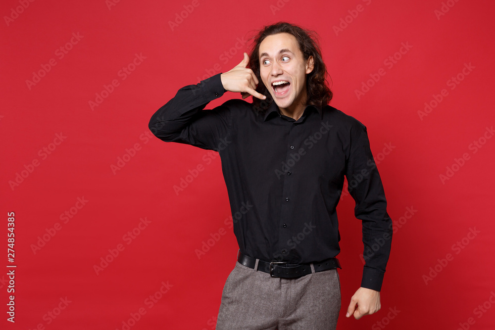 Young curly long haired man in black shirt posing isolated on red wall background studio portrait. People emotions lifestyle concept. Mock up copy space. doing phone gesture like says call me back.