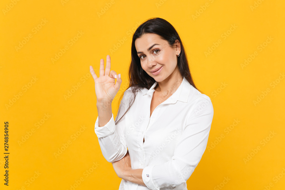 Portrait of smiling confident young woman in white shirt showing OK gesture, looking camera isolated on bright yellow orange wall background in studio. People lifestyle concept. Mock up copy space. Portrait of smiling confident young woman in white shirt showing OK gesture, looking camera isolated on bright yellow orange wall background in studio. People lifestyle concept. Mock up copy space.