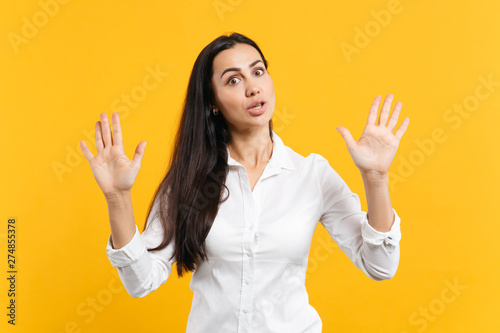Portrait of shocked young woman in white shirt looking camera, rising hands, showing palms isolated on bright yellow orange wall background in studio. People lifestyle concept. Mock up copy space.