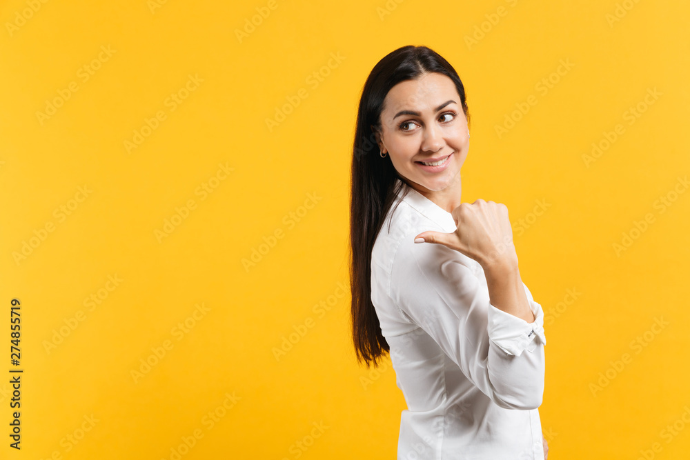 Side view of beautiful smiling young woman in white casual shirt pointing thumb aside behind her back isolated on yellow orange wall background in studio. People lifestyle concept. Mock up copy space. Side view of beautiful smiling young woman in white casual shirt pointing thumb aside behind her back isolated on yellow orange wall background in studio. People lifestyle concept. Mock up copy space.