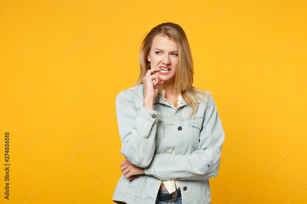 Portrait of preoccupied nervous young woman in denim casual clothes looking aside, gnawing nails isolated on yellow orange wall background in studio. People lifestyle concept. Mock up copy space. Portrait of preoccupied nervous young woman in denim casual clothes looking aside, gnawing nails isolated on yellow orange wall background in studio. People lifestyle concept. Mock up copy space.