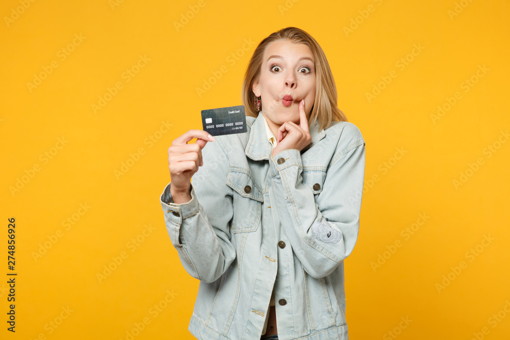 Portrait of surprised amazed shocked young woman in denim casual clothes holding credit bank card isolated on yellow orange wall background in studio. People lifestyle concept. Mock up copy space. Portrait of surprised amazed shocked young woman in denim casual clothes holding credit bank card isolated on yellow orange wall background in studio. People lifestyle concept. Mock up copy space.