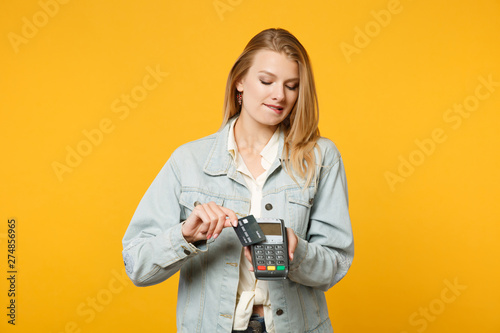 Young woman biting lips, holding wireless modern bank payment terminal to process and acquire credit card payments isolated on yellow orange background. People lifestyle concept. Mock up copy space.