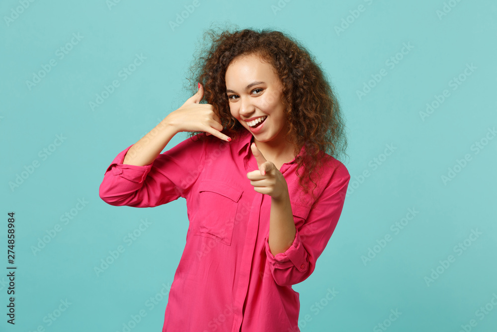 Smiling african girl doing phone gesture like says call me back, pointing index finger on camera isolated on blue turquoise background. People sincere emotions, lifestyle concept. Mock up copy space.