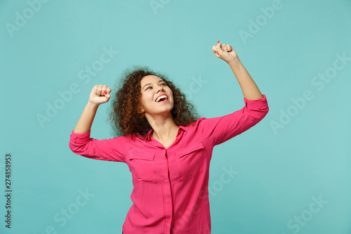 Portrait of cheerful african girl in casual clothes doing winner gesture looking up isolated on blue turquoise wall background in studio. People sincere emotions lifestyle concept. Mock up copy space. © ViDi Studio