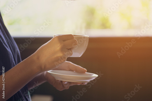 Beautiful young asian women hands hold a warm white cup of coffee standing next to a window with sunlight. Close up portrait of Asian woman with a cup of morning coffee in hands.