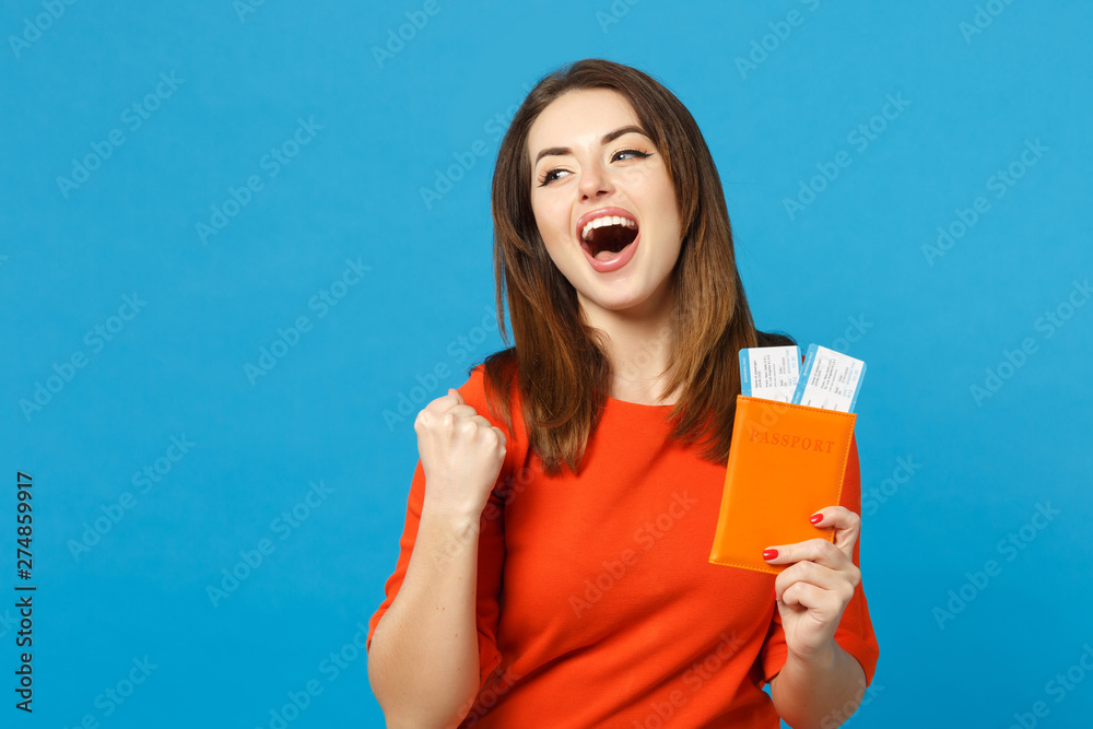 Beautiful brunette young woman wearing red orange dress hol in hand passport tickets isolated over trendy blue wall background, studio portrait. People lifestyle fashion concept. Mock up copy space.