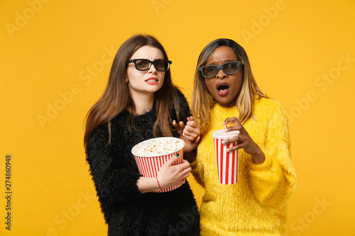 Two women friends european and african american in black yellow clothes hold bucket of popcorn isolated on bright orange wall background, studio portrait. People lifestyle concept. Mock up copy space.
