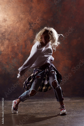 Photo of long-haired blonde dancer woman with developing hair in torn jeans and sneakers on dark brown background