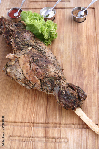 Big delicious baked leg of lamb. With greens and sauces. On a wooden board. On a white background.