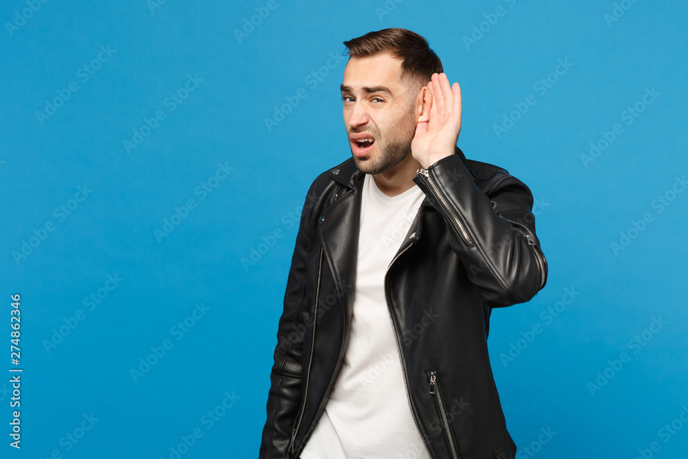 Young sad frustrated worried unshaven man in black jacket white t-shirt try to hear you isolated on blue wall background studio portrait. People sincere emotions lifestyle concept. Mock up copy space. Young sad frustrated worried unshaven man in black jacket white t-shirt try to hear you isolated on blue wall background studio portrait. People sincere emotions lifestyle concept. Mock up copy space.