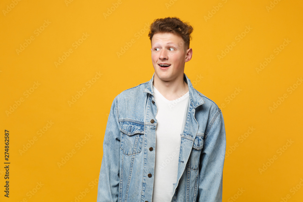 Portrait of perplexed young man in denim casual clothes looking aside keeping mouth open isolated on yellow orange background in studio. People sincere emotions, lifestyle concept. Mock up copy space. Portrait of perplexed young man in denim casual clothes looking aside keeping mouth open isolated on yellow orange background in studio. People sincere emotions, lifestyle concept. Mock up copy space.