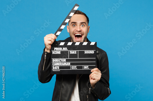 Handsome stylish young unshaven man in black jacket white t-shirt hold in hand film making clapperboard isolated on blue wall background studio portrait. People lifestyle concept. Mock up copy space. © ViDi Studio