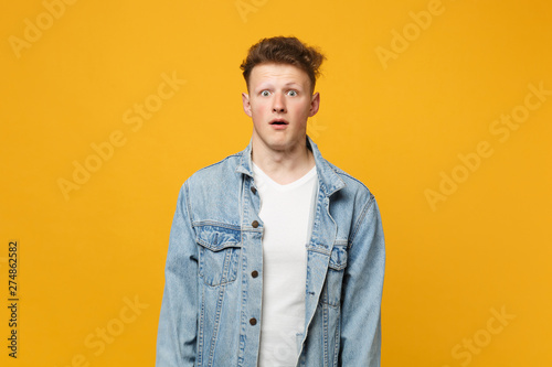 Portrait of shocked young man in denim casual clothes looking camera, keeping mouth open isolated on yellow orange background in studio. People sincere emotions, lifestyle concept. Mock up copy space.