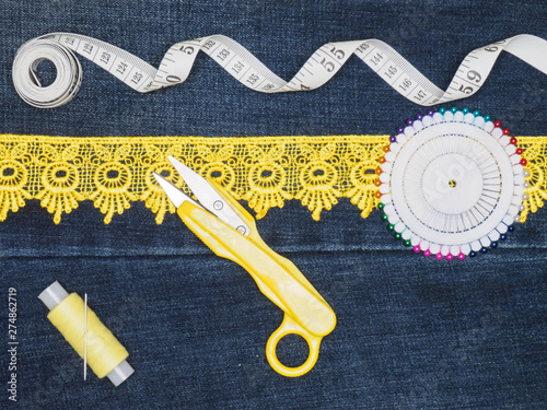 Yellow lace, thread with a needle, a tape measure, pins, scissors on a jeans background. Sewing still life photo