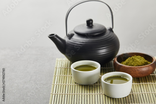 selective focus of green matcha powder in wooden bowl near black teapot and cups with tea on bamboo table mat