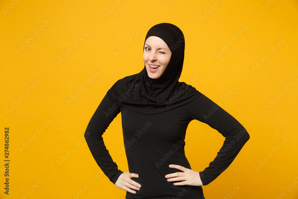 Young arabian muslim woman in hijab black clothes standing with arms akimbo on waist isolated on yellow wall background, studio portrait. People religious Islam lifestyle concept. Mock up copy space. Young arabian muslim woman in hijab black clothes standing with arms akimbo on waist isolated on yellow wall background, studio portrait. People religious Islam lifestyle concept. Mock up copy space.