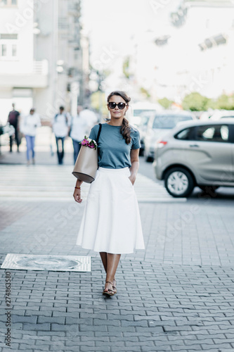 Fashionable young woman in sunglasses, a white skirt and a blue blouse walks city with a bag and flowers. European retro style.