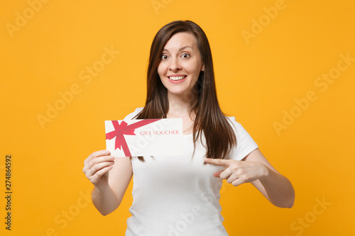 Portrait of pretty young woman in white casual clothes pointing index finger on gift certificate isolated on bright yellow orange background in studio. People lifestyle concept. Mock up copy space.