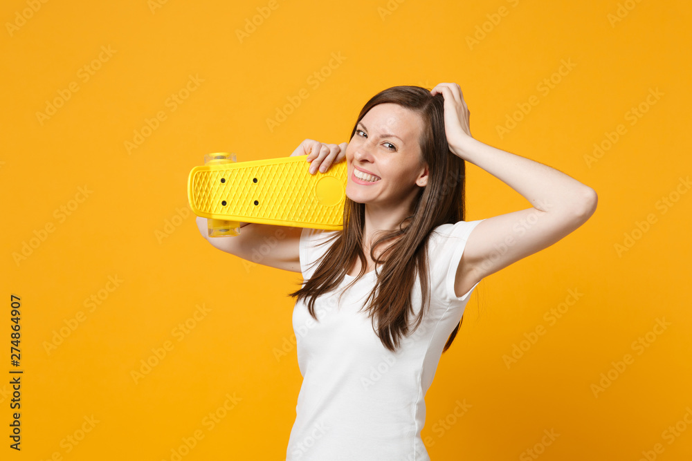 Portrait of smiling young woman in white casual clothes putting hand on head, holding yellow skateboard isolated on bright yellow orange wall background. People lifestyle concept. Mock up copy space. Portrait of smiling young woman in white casual clothes putting hand on head, holding yellow skateboard isolated on bright yellow orange wall background. People lifestyle concept. Mock up copy space.