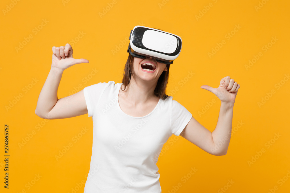 Laughing young girl in white casual clothes watching in headset of virtual reality pointing thumbs on herself isolated on bright yellow orange background. People lifestyle concept. Mock up copy space. Laughing young girl in white casual clothes watching in headset of virtual reality pointing thumbs on herself isolated on bright yellow orange background. People lifestyle concept. Mock up copy space.
