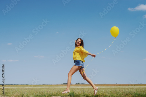 Gorgeous Caucasian teenage girl in yellow blouse, denim shorts and with eyeglasses running outdoor with yellow balloon in hand.