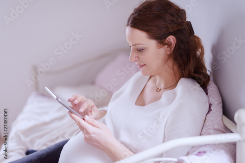 Beautiful Caucasian pregnant woman sitting on sofa in living room and using tablet.