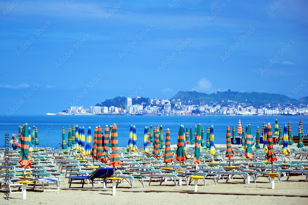 Closed umbrellas and empty chaise lounges at the sandy beach. Beach chairs, beds and umbrellas on the Albanian beach before summer holiday season. Golem, Durres, Albania. Durres city in a distance