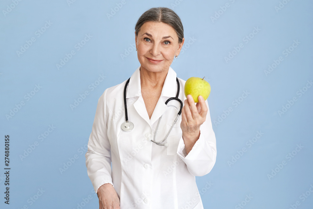 Isolated image of confident friendly looking mature senior woman dentist offering you green apple for dental care. Attractive female doctor wearing stethoscope recommending eating more fruits Isolated image of confident friendly looking mature senior woman dentist offering you green apple for dental care. Attractive female doctor wearing stethoscope recommending eating more fruits