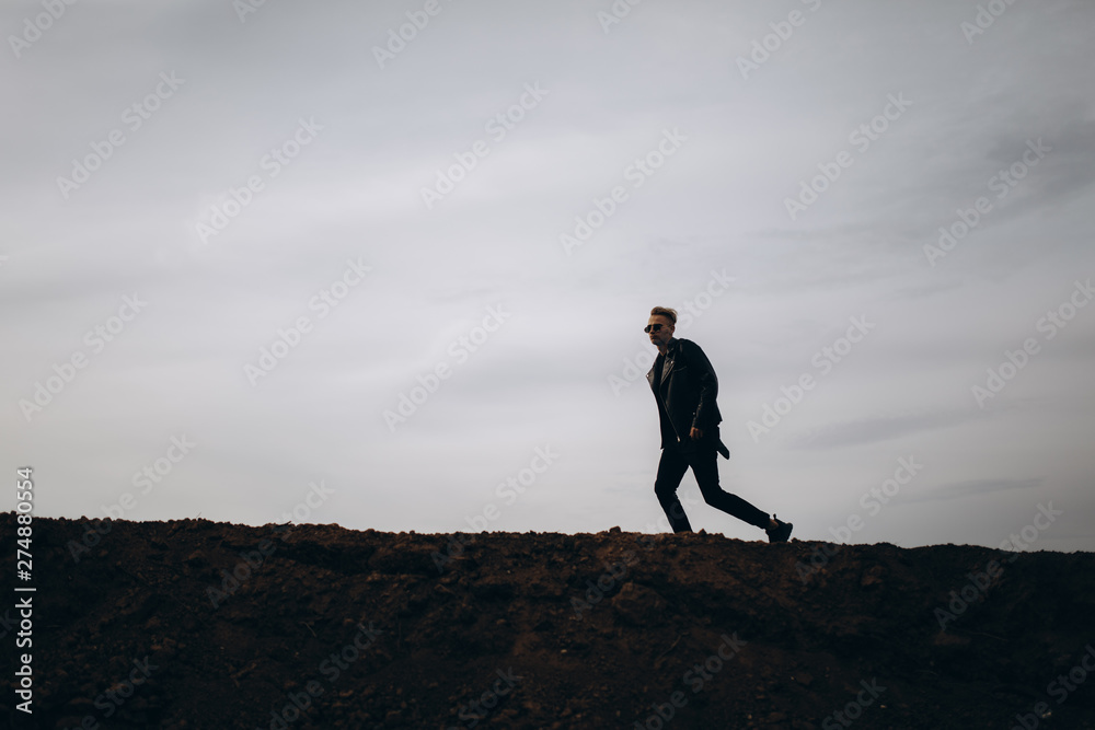 Young sexy running man in leather jacket and sunglasses standing outdoor. Silhouette against grey sky Young sexy running man in leather jacket and sunglasses standing outdoor. Silhouette against grey sky