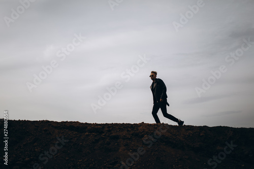 Young sexy running man in leather jacket and sunglasses standing outdoor. Silhouette against grey sky