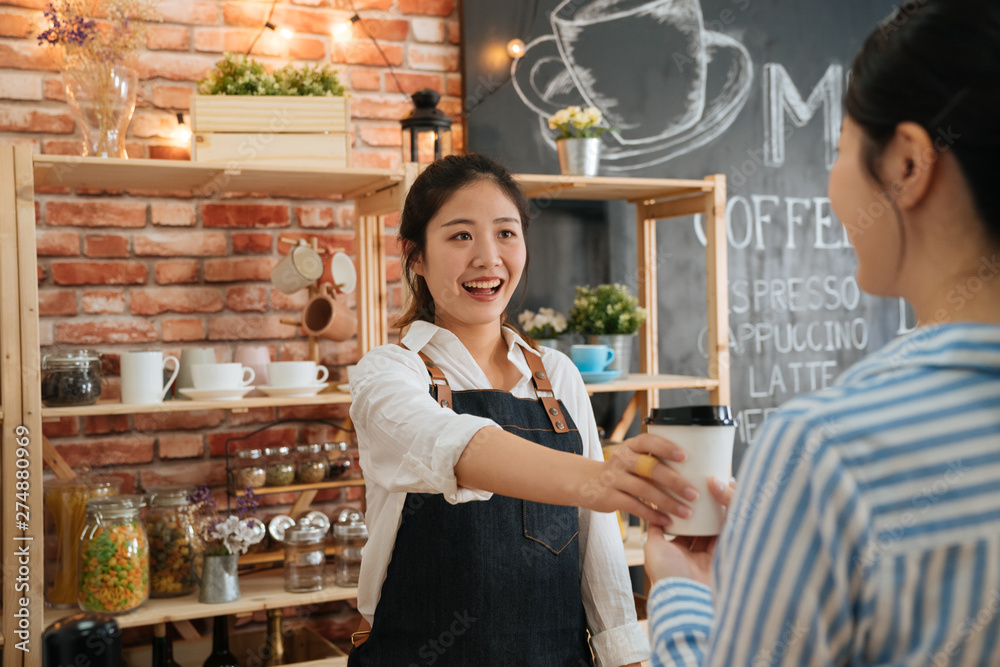 Beautiful friendly female barista giving coffee disposable cup to customer at counter of coffee