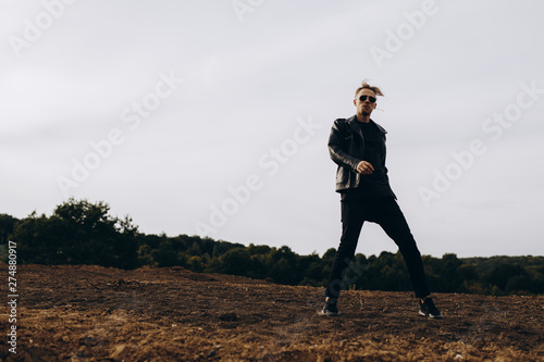 Young sexy man in leather jacket and sunglasses standing outdoor. Silhouette against grey sky