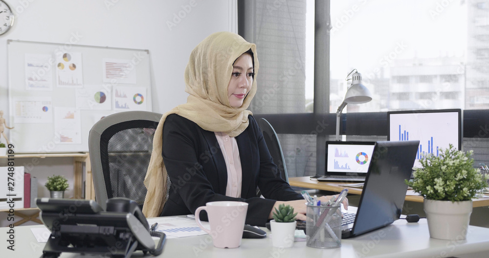 Young islam female employee looking at computer monitor during working day in office typing on laptop keyboard. elegant positive muslim lady manager in suit sitting workplace desk with morning coffee Young islam female employee looking at computer monitor during working day in office typing on laptop keyboard. elegant positive muslim lady manager in suit sitting workplace desk with morning coffee