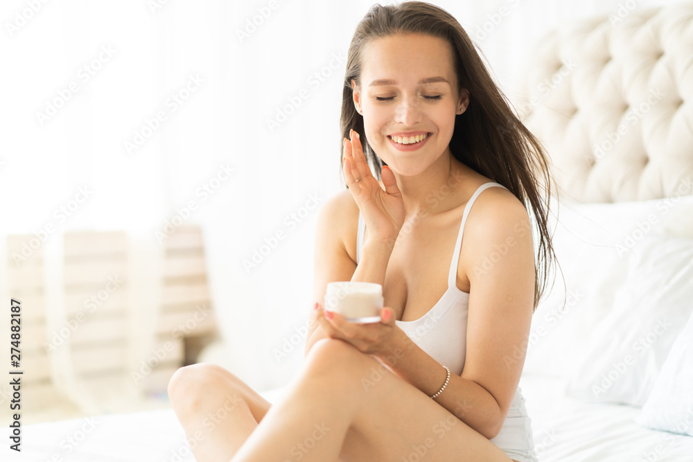 Pretty brunette using body cream on bed at home. Pretty brunette using body cream on bed at home.