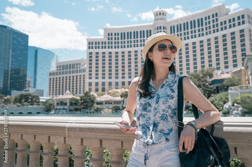 laughing girl traveler in summer attire leaning on railing and reading mobile messages with big fountain near famous hotel behind. Outdoor of woman in sunglasses chilling on city square in sunny day.