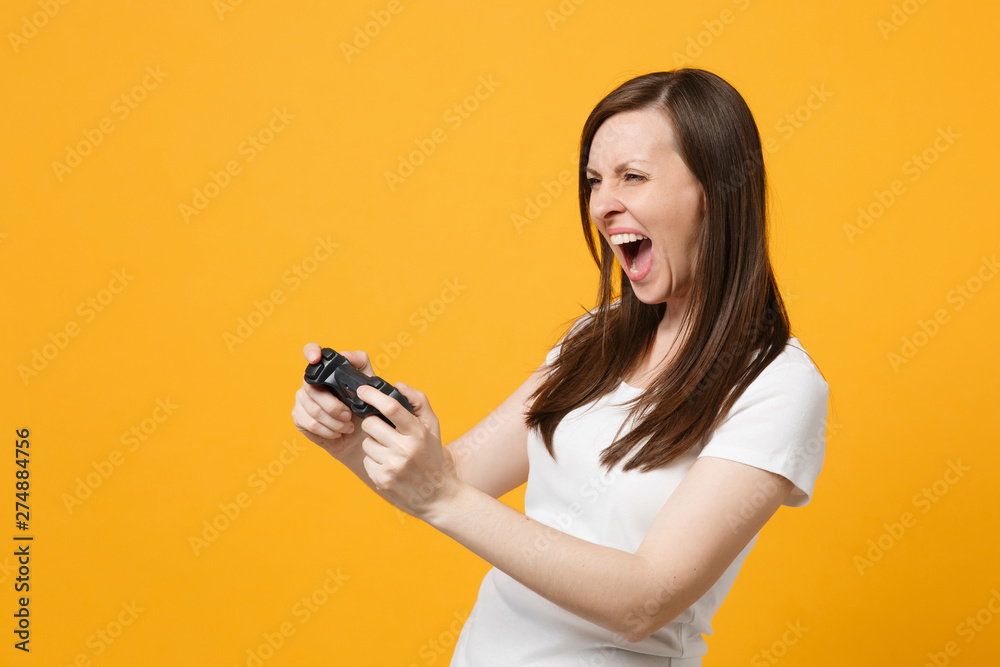 Side view of screaming young woman in white casual clothes playing video game with joystick isolated on bright yellow orange wall background in studio. People lifestyle concept. Mock up copy space. Side view of screaming young woman in white casual clothes playing video game with joystick isolated on bright yellow orange wall background in studio. People lifestyle concept. Mock up copy space.
