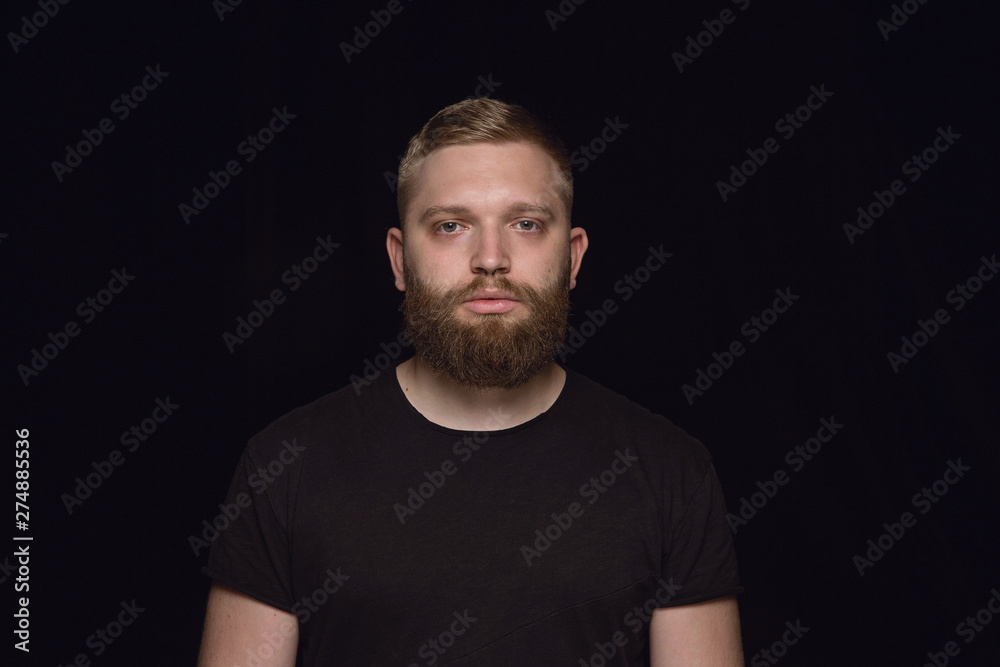 Close up portrait of young caucasian man isolated on black studio background. Photoshot of real emotions of male model. Standing and looks serious. Facial expression, human nature and emotions concept