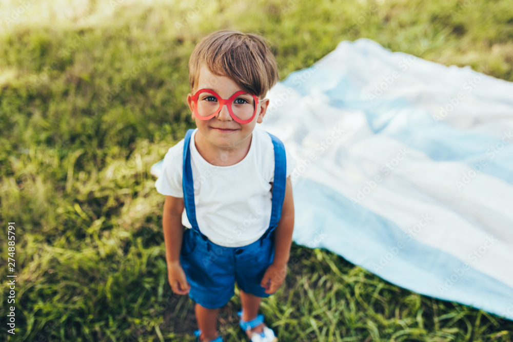 Top image of funny little boy wearing blue shorts and red glasses playing at nature background. Cheerful child posing on the green grass in the park. Kid having fun on sunlight outdoors. Childhood. Top image of funny little boy wearing blue shorts and red glasses playing at nature background. Cheerful child posing on the green grass in the park. Kid having fun on sunlight outdoors. Childhood.