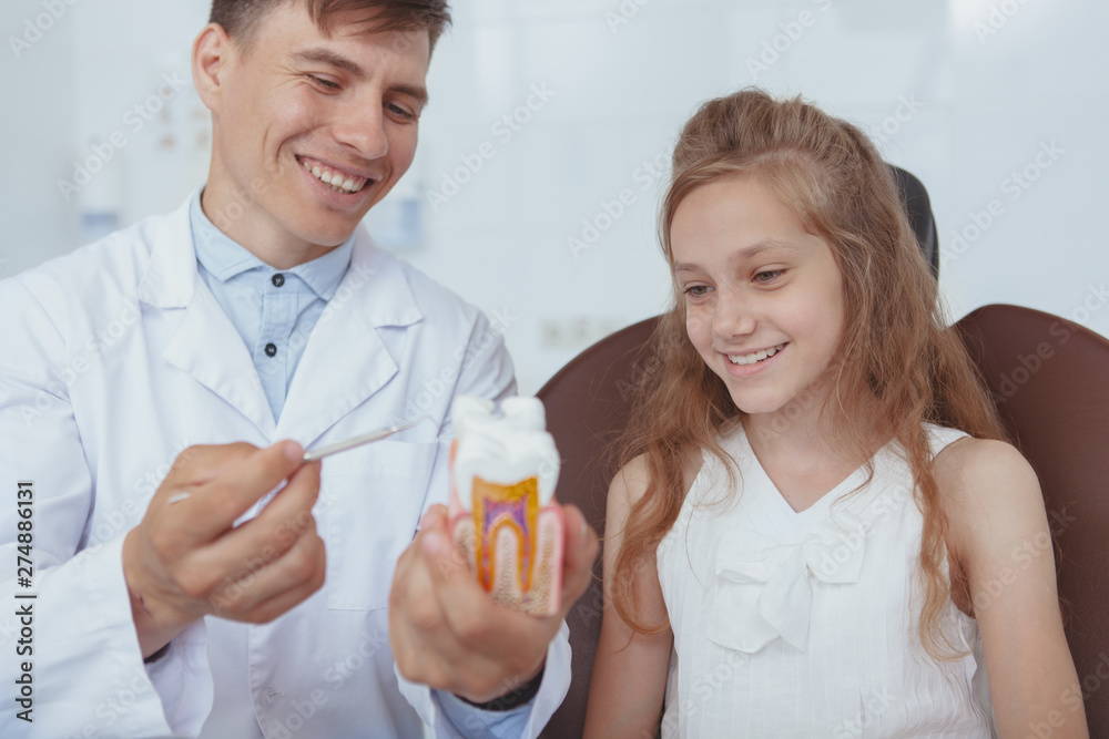 Cheerful young male dentist showing tooth model to his young patient. Lovely girl learning about dental care during medical appointment with dentist Cheerful young male dentist showing tooth model to his young patient. Lovely girl learning about dental care during medical appointment with dentist