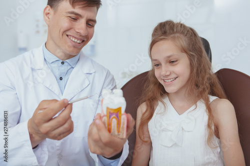 Cheerful young male dentist showing tooth model to his young patient. Lovely girl learning about dental care during medical appointment with dentist